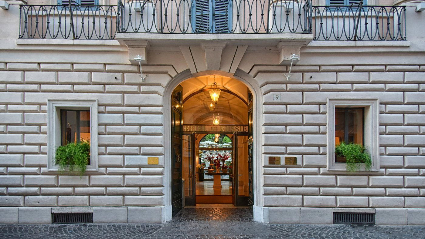 Elegant entrance to Hotel de Russie with stone facade, arched doorway, and warm lights. Potted plants decorate windows, creating a cozy feel.
