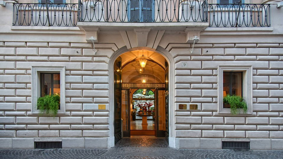 Elegant entrance to Hotel de Russie with stone facade, arched doorway, and warm lights. Potted plants decorate windows, creating a cozy feel.