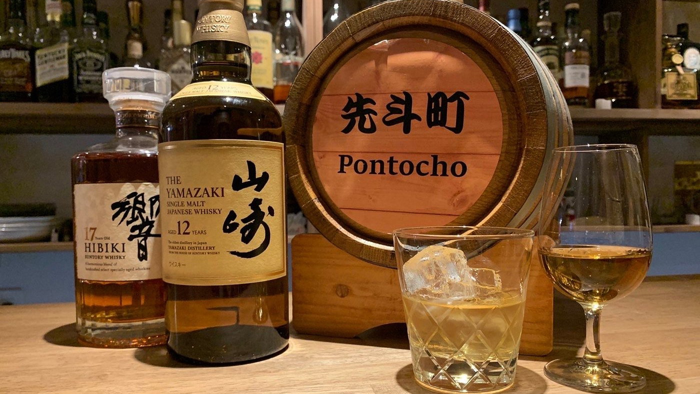 Bottles of Hibiki and Yamazaki whiskey on a bar, with a glass of whiskey and ice, a wooden barrel with "Pontocho," and shelves in the background.
