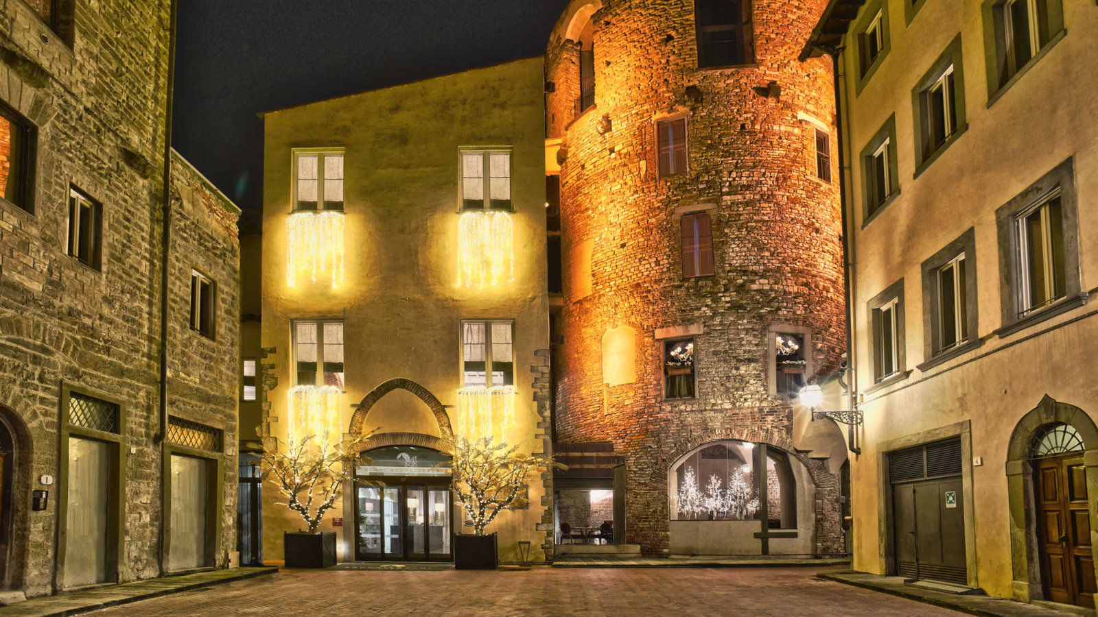 Historic buildings at night with warm golden lights, festive decorations, and illuminated trees in a quiet cobblestone street setting.