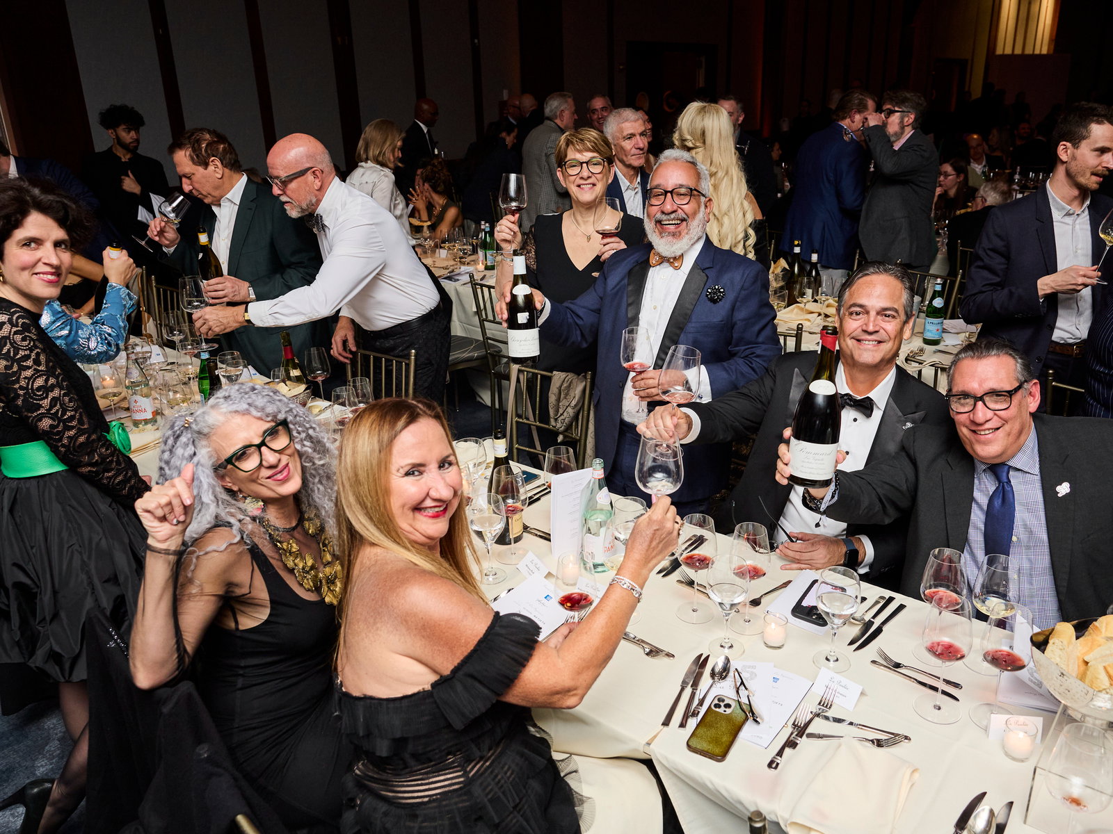 Revelers at the 2025 La Paulée NYC Gala Dinner in formal attire toast with wine. Tables are set with glasses, menus, and candles. Joyful atmosphere.