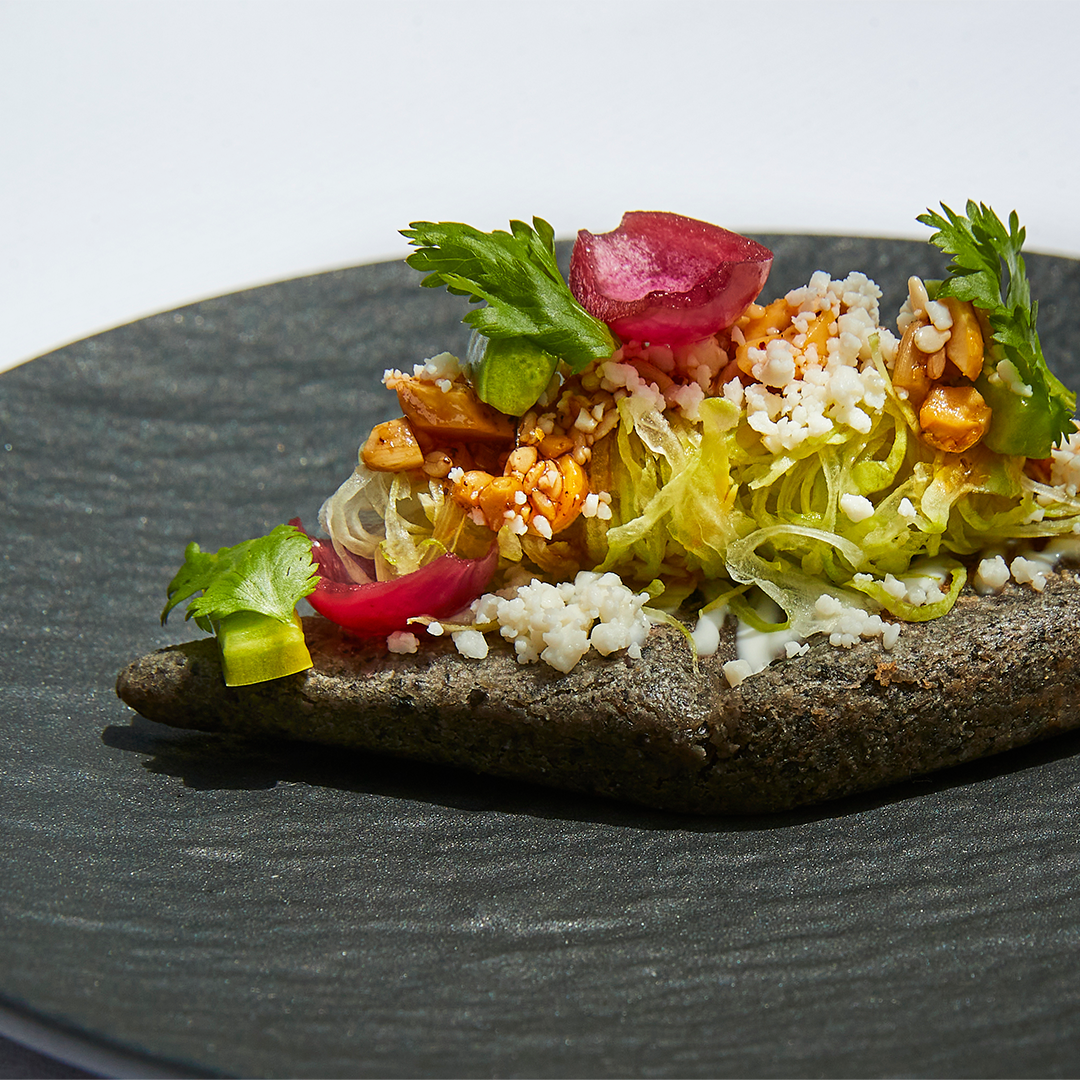 Gourmet taco on a black plate, topped with lettuce, nuts, pink radish, and crumbled cheese, garnished with green cilantro.