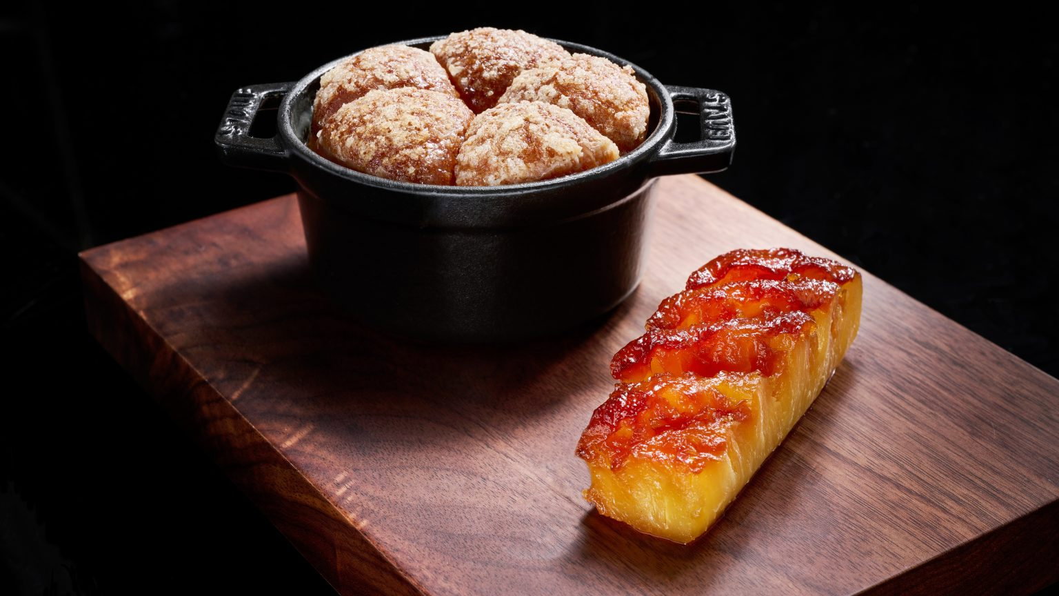 Rustic black pot with sugared buns next to caramelized pineapple slice on wooden board, against a dark background. Warm, inviting scene.