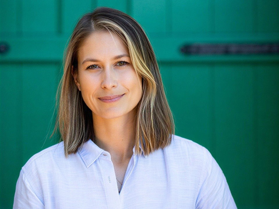 Winemaker Maya Dalla Valle with a warm smile stands against a bright green door, wearing a white shirt. The mood is calm and friendly.