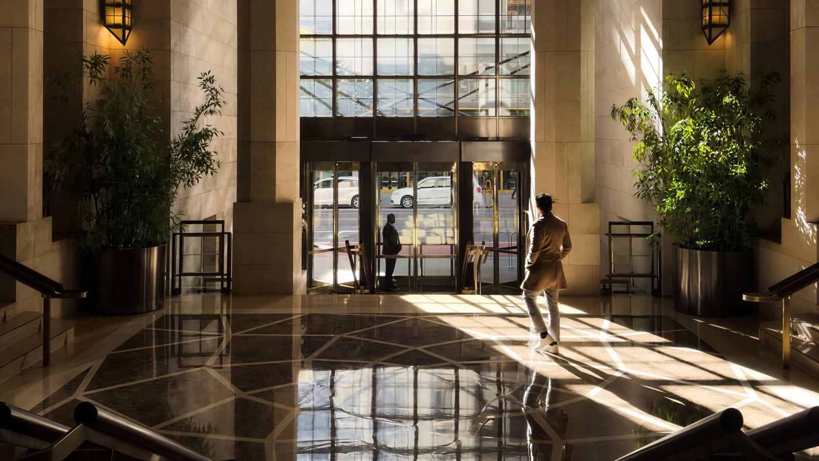 Man in a coat walks through a sunlit, marble-floored lobby with large glass doors, green plants, and geometric patterns, creating a calm mood.
