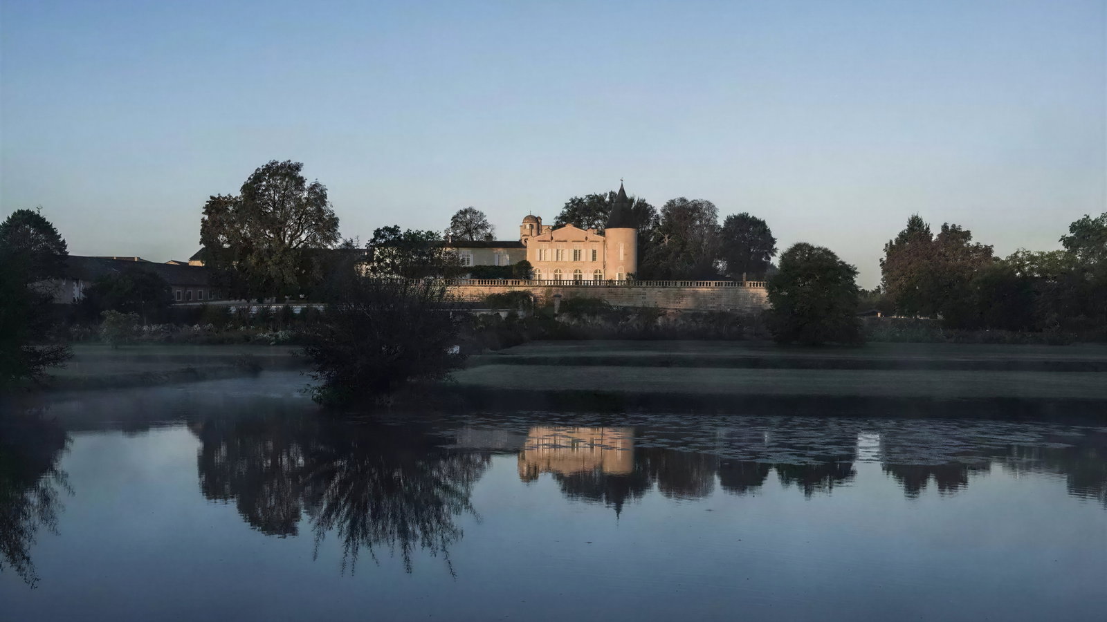 A historic mansion with a turret reflects on a calm river, surrounded by trees at dusk. The sky is clear, creating a serene mood.