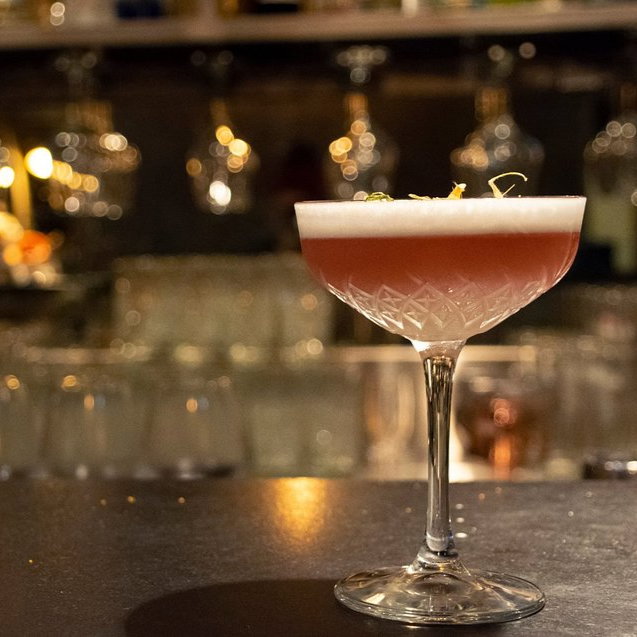 Elegant cocktail in a crystal glass on a bar counter, with a frothy top and warm lighting in the background. Mood is classy and relaxed.