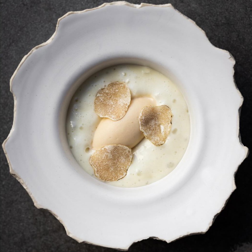 A white bowl with cream, topped with three truffle slices and a dollop of beige puree. Gray textured background, elegant and minimalistic.