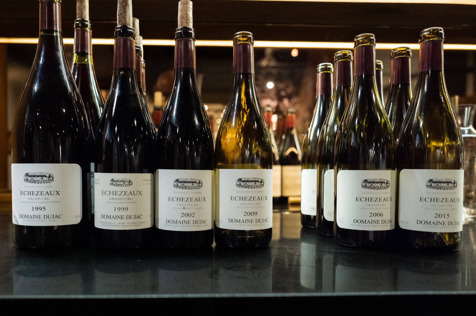 Wine bottles labeled "Echezeaux Grand Cru, Domaine Dujac" from various years are lined up on a reflective surface. Warm lighting.