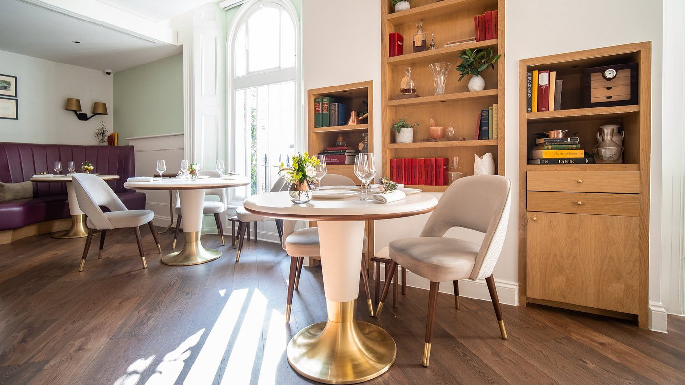 Bright dining area with round tables, elegant gray chairs, gold accents, and bookshelves. Sunlit wooden floor adds a cozy ambiance.
