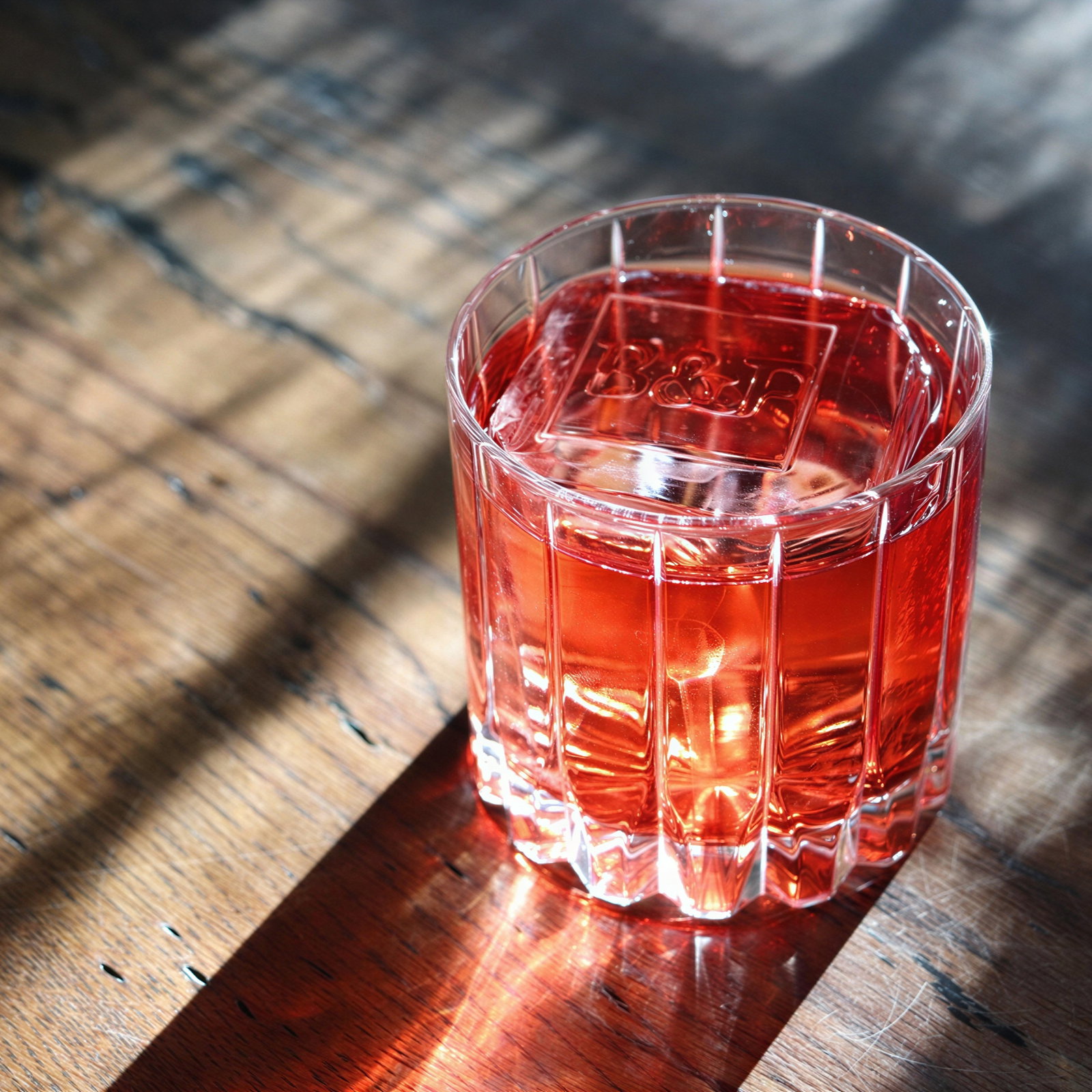 A glass of red liquid with ice sits on a wooden table, casting shadows. Sunlight highlights the drink's vibrant color.
