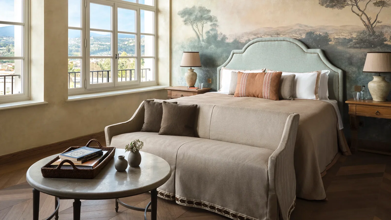 Grand suite bedroom at Collegio alla Querce with a large bed and a fresco painted behind it. There is a beige sofa, and round table holding a tray and vases. Mural with trees, large windows with scenic view.