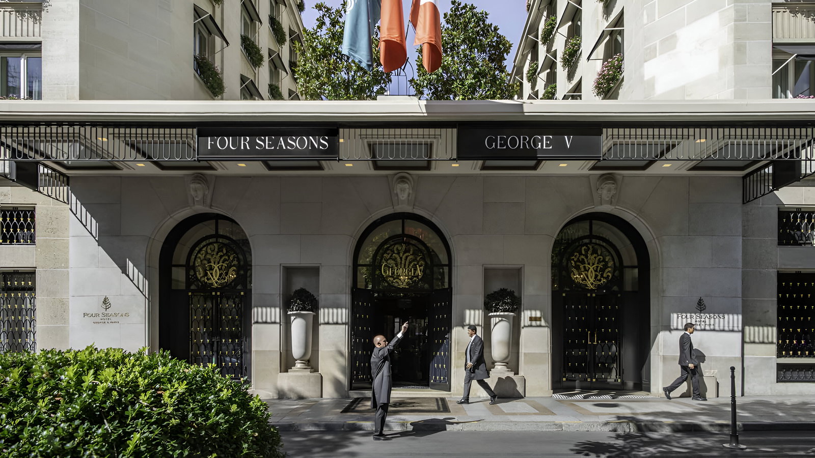 Facade of Four Seasons George V hotel with two men in suits walking, one raising his hand. Black, white, and green elements, flags above.