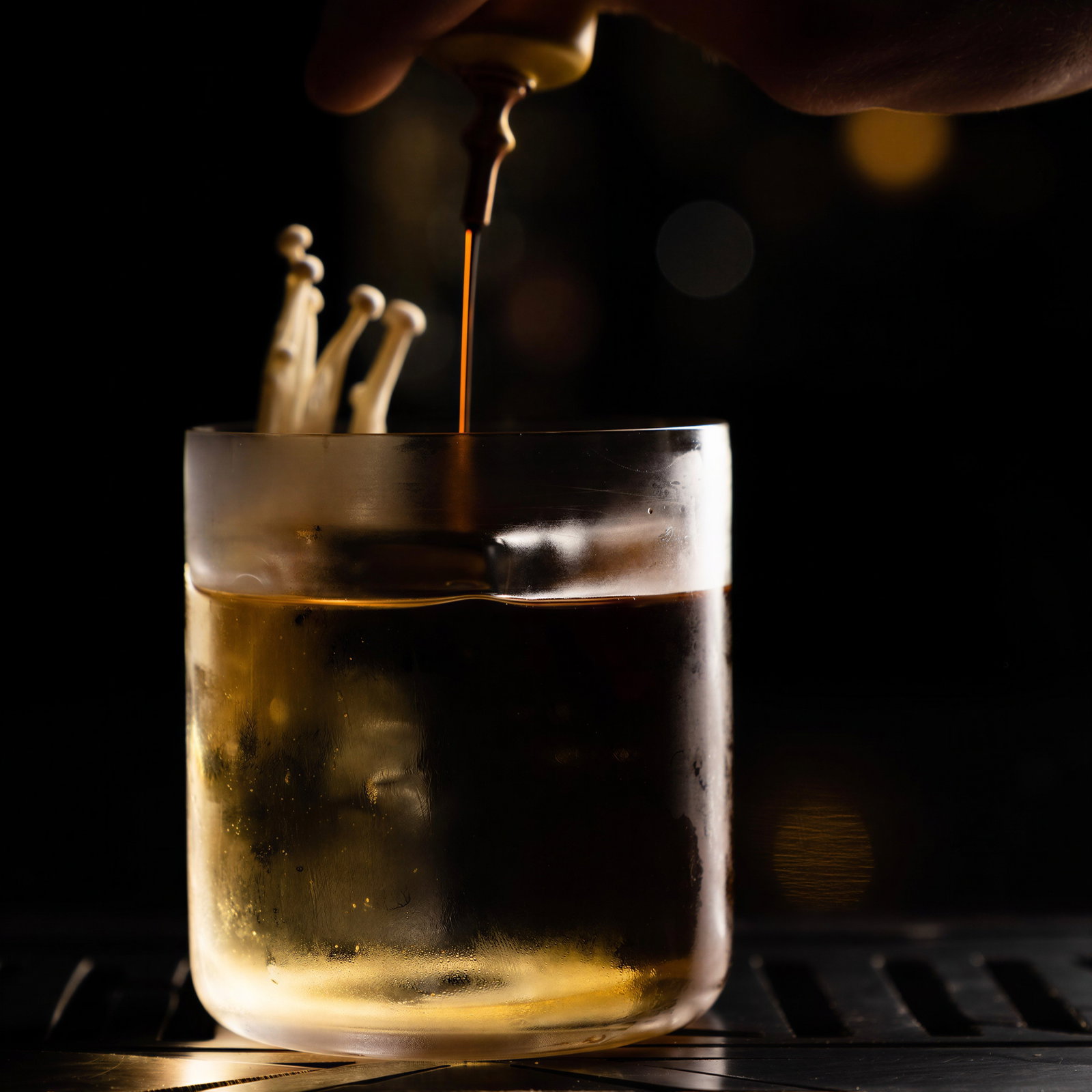 A hand pours liquid from a dropper into a glass with mushrooms, set against a dark background with warm lighting.