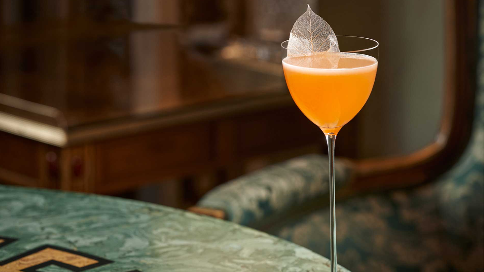 Elegant cocktail in a coupe glass with an orange liquid and leaf garnish on a green marble table. Classic wooden furniture in the background.