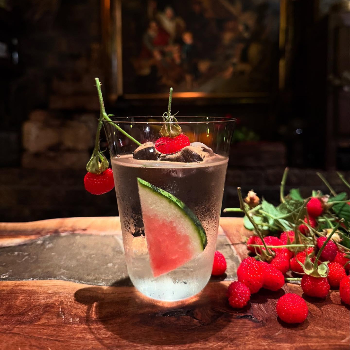 Frosted glass with ice, raspberry, and watermelon slice on a wood table, surrounded by fresh raspberries; dim, rustic setting.