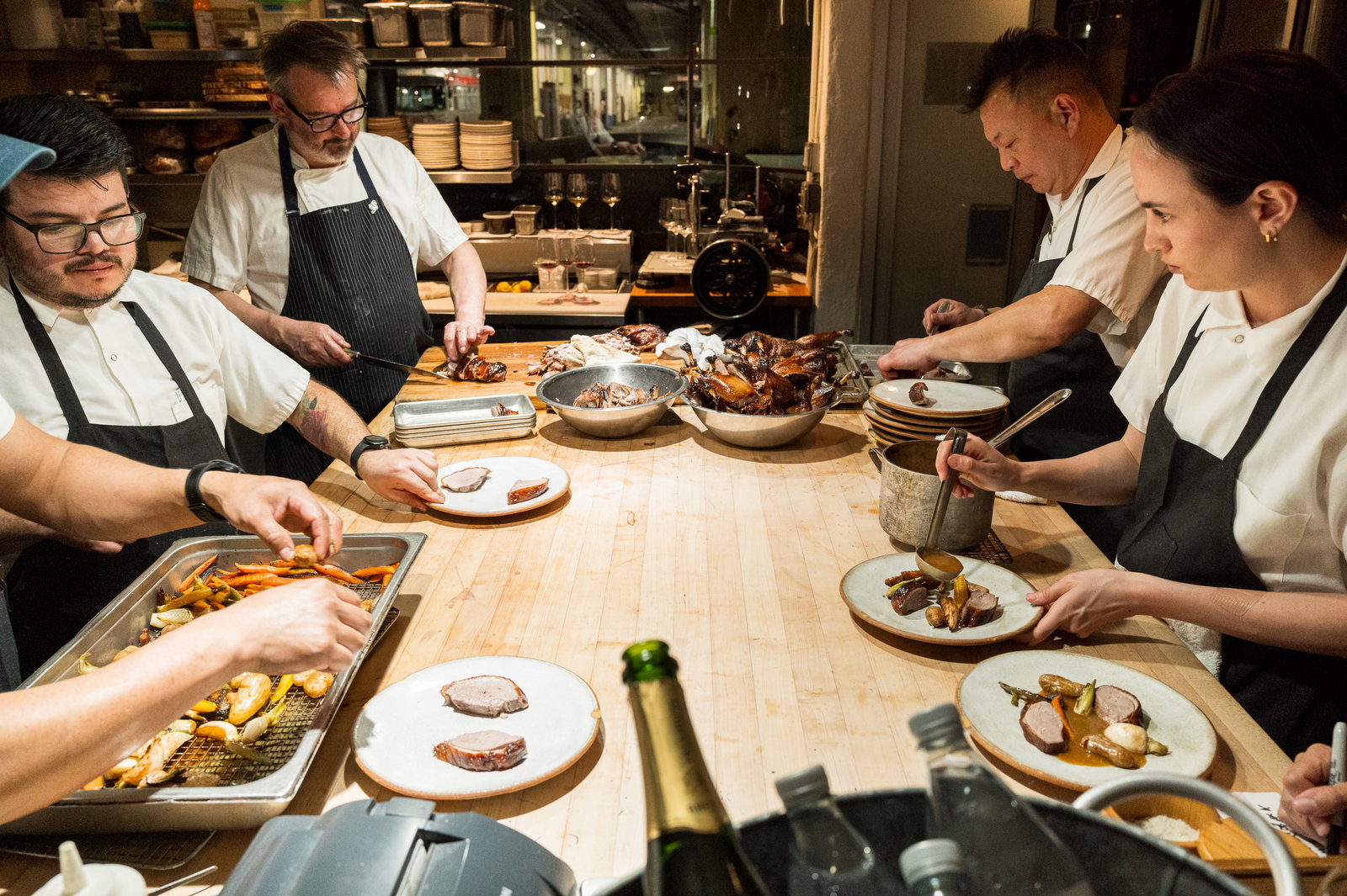 Chefs in white shirts and black aprons prepare dishes at a wooden kitchen counter, surrounded by plates and food, creating a focused atmosphere.