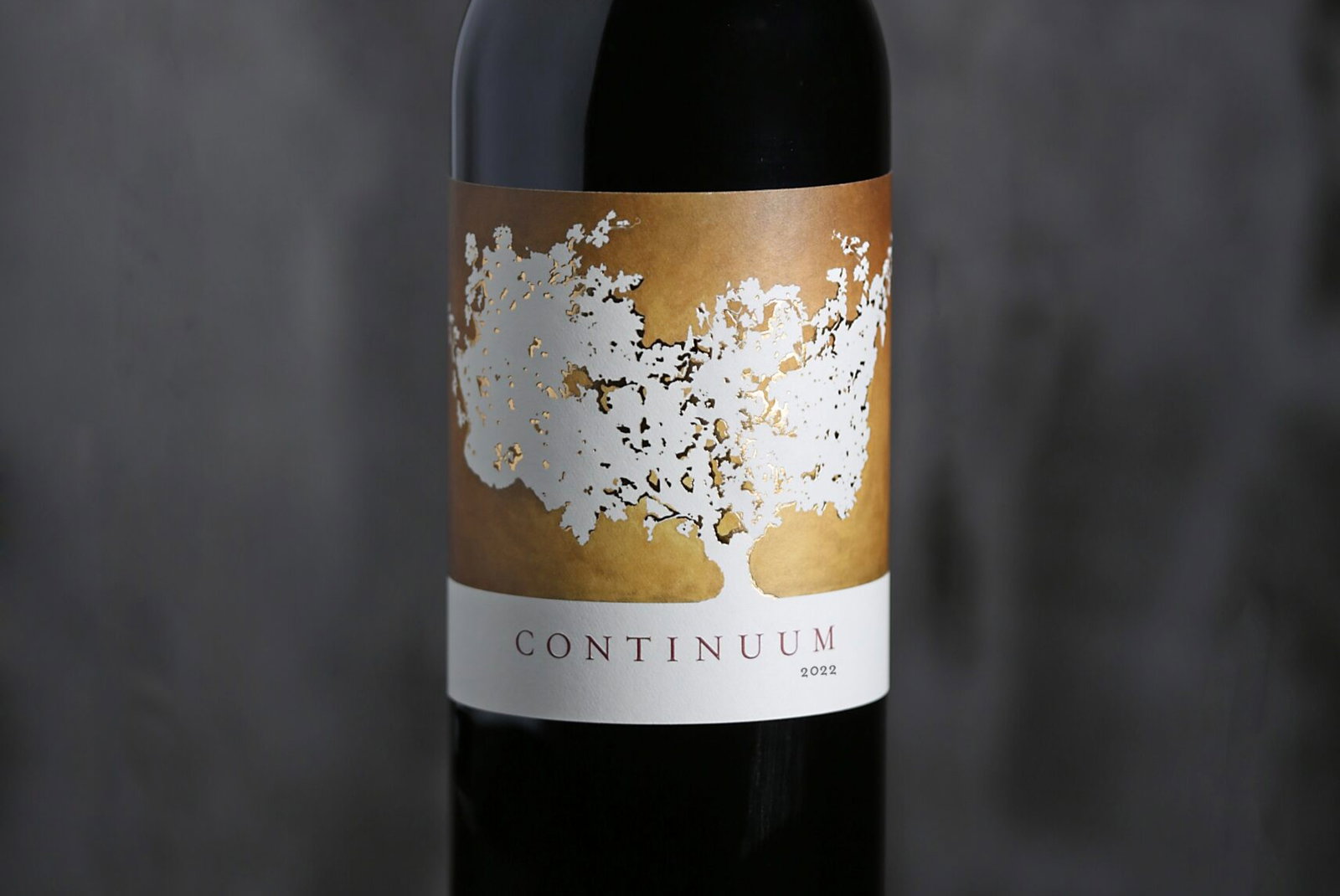 Wine bottle with a brown label featuring white tree artwork. Text reads "Continuum 2022." Background is dark and blurred.