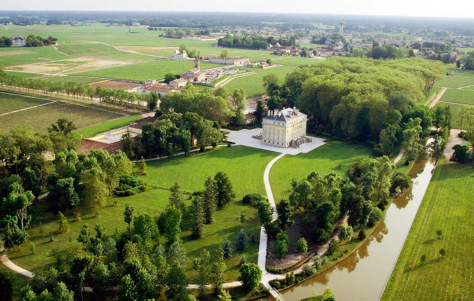 Aerial view of a grand estate with a large mansion, vibrant green lawns, surrounding trees, and a winding river, set in a rural landscape.