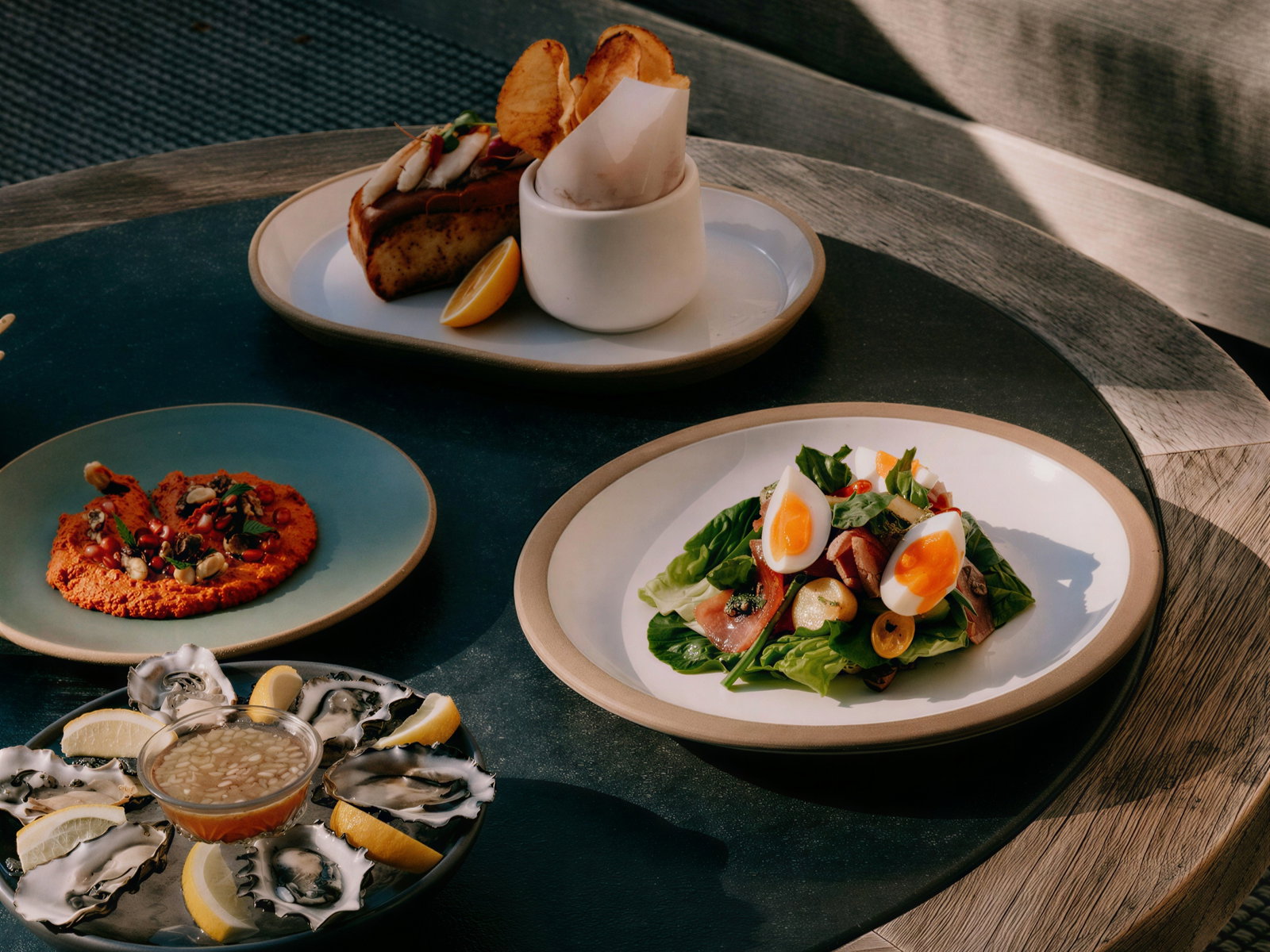 A table with various dishes: oysters with lemon, a salad with eggs, hummus, and a sandwich. Set outdoors on a wooden table.