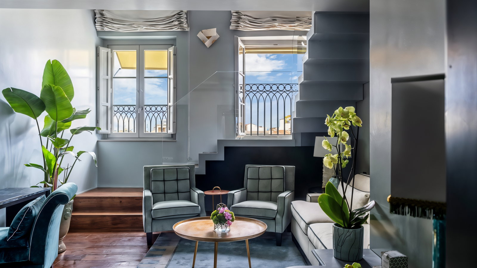 Elegant living room with green chairs, a round wooden table, and potted plants. Two open windows with views of blue sky, neutral tones.