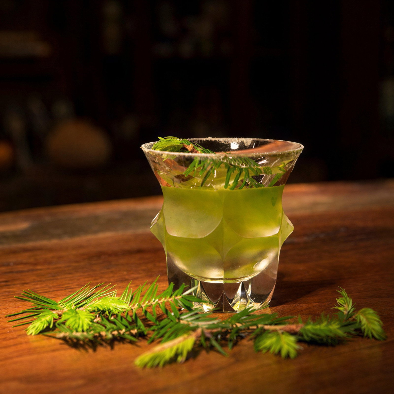 Green cocktail in a textured glass with pine sprigs, set on a wooden table. Dark, blurred background creates a cozy, rustic atmosphere.