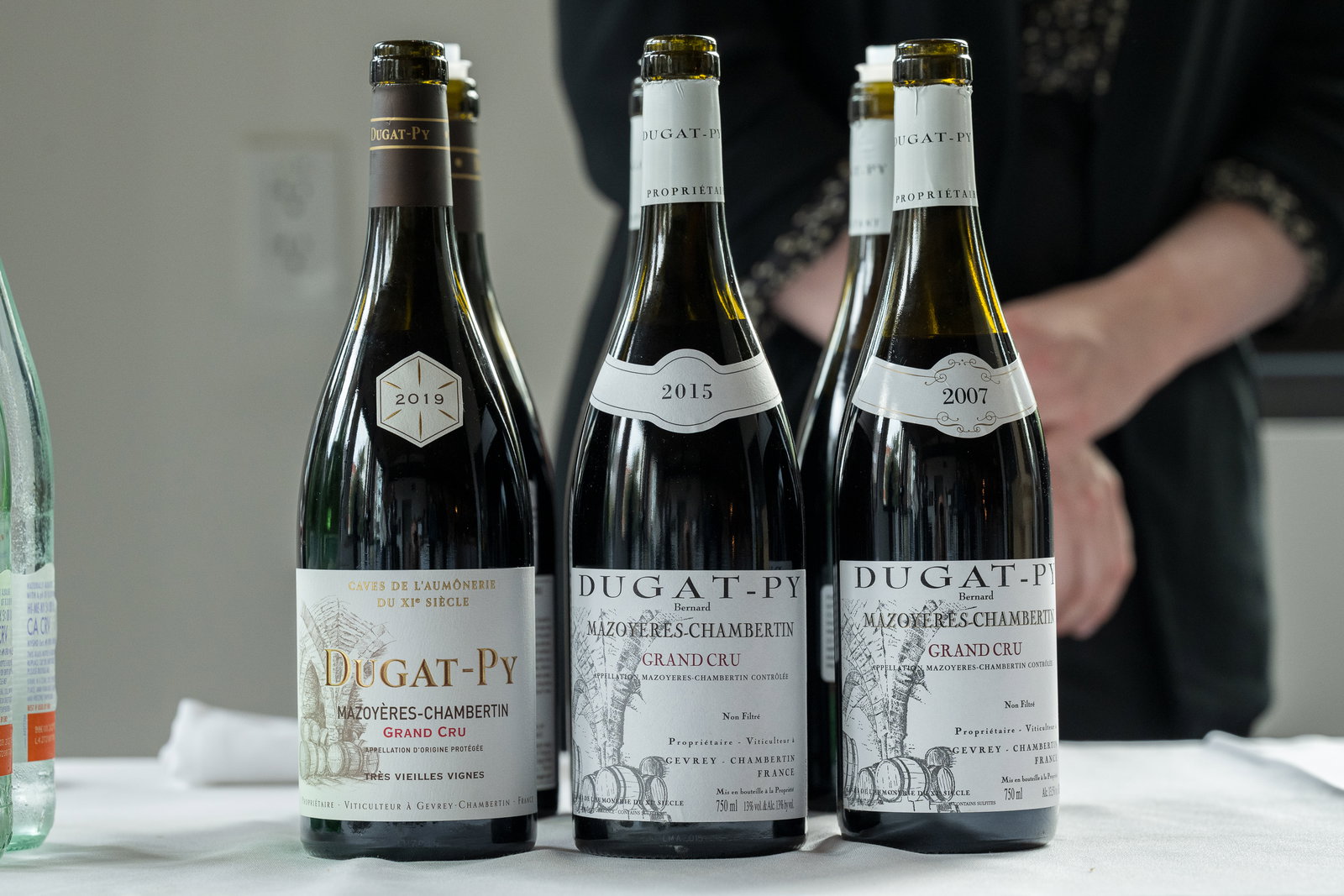 Three Dugat-Py wine bottles (2019, 2015, 2007) on a white tablecloth, with a person's hands in the background. Elegant and moody setting.