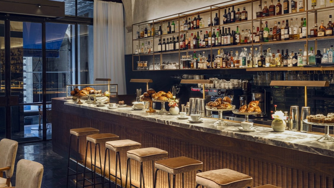 Modern bar interior with a marble counter displaying pastries and coffee cups. Shelves with liquor bottles adorn the wall. Warm lighting.