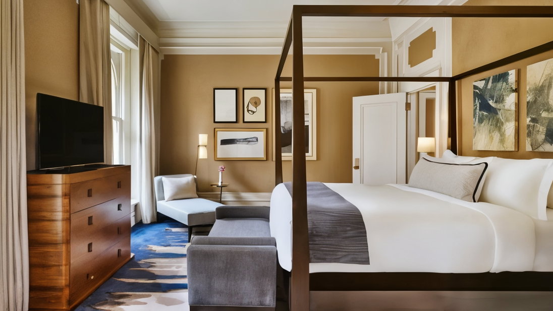 Elegant hotel room with a four-poster bed, modern art, a wooden dresser, and a chaise lounge. Neutral tones, blue carpet, cozy ambiance.