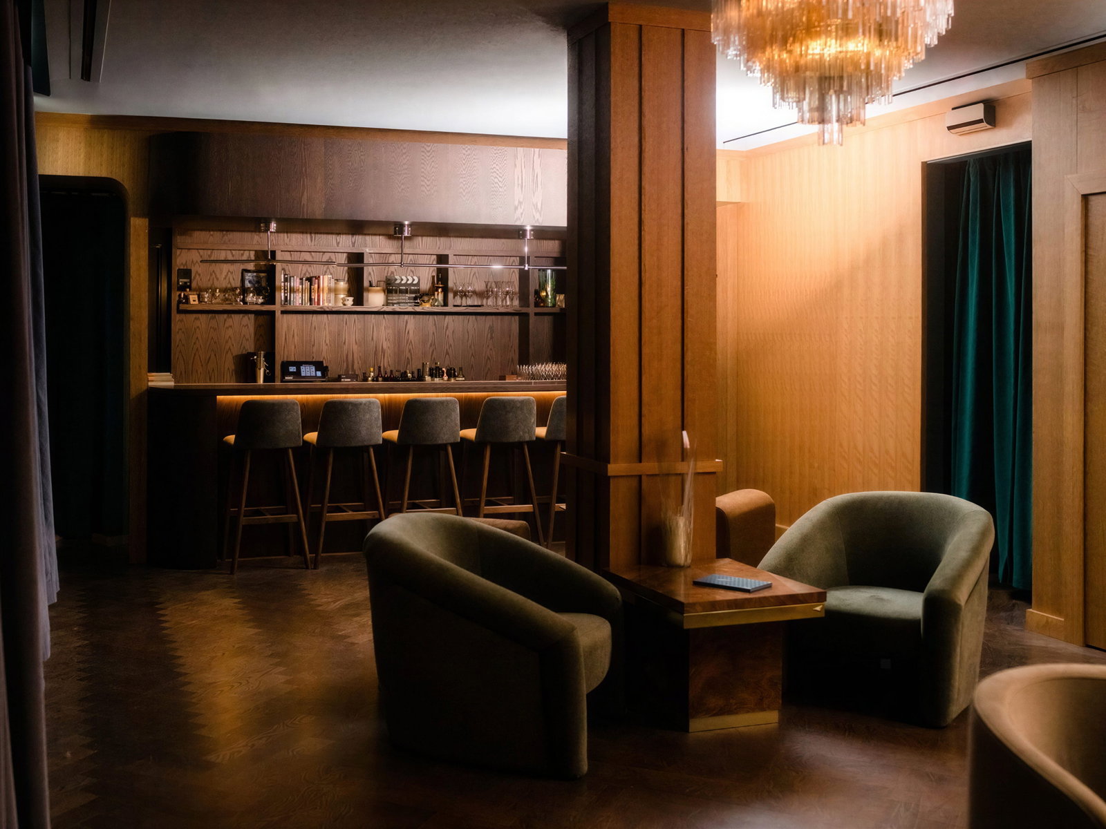 Cozy lounge with empty green chairs, wooden table, and modern bar in warm lighting. A chandelier hangs from the ceiling.