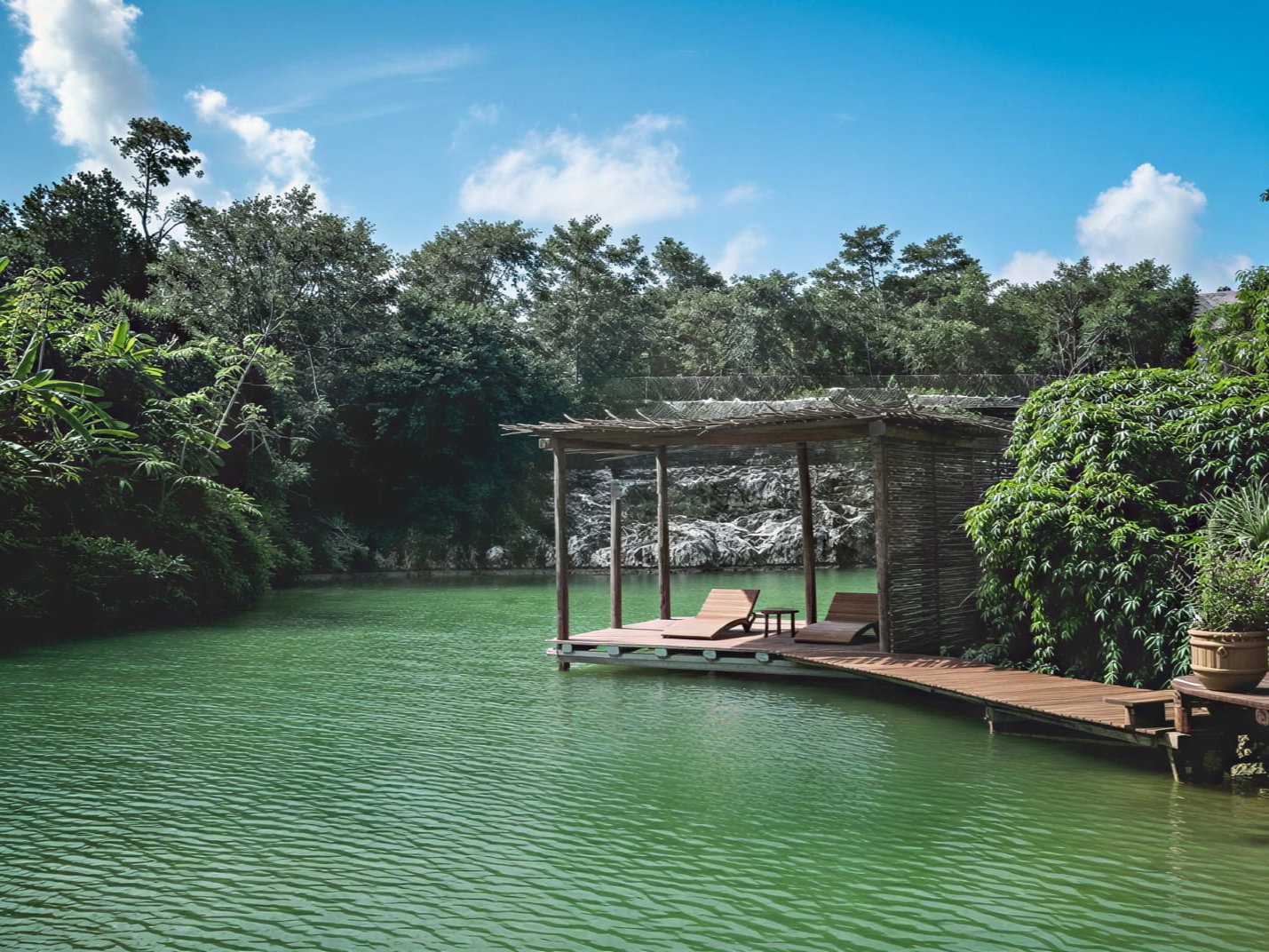 A tranquil lake cenote at Wakax Hacienda with a shaded wooden deck featuring two lounge chairs. Surrounded by lush green trees under a clear blue sky.