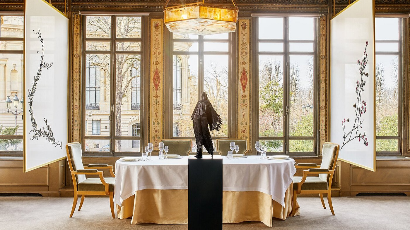 Elegant dining room with large windows and tree views. A round table with white cloth, set for dining, features a center statue. Art panels are present.