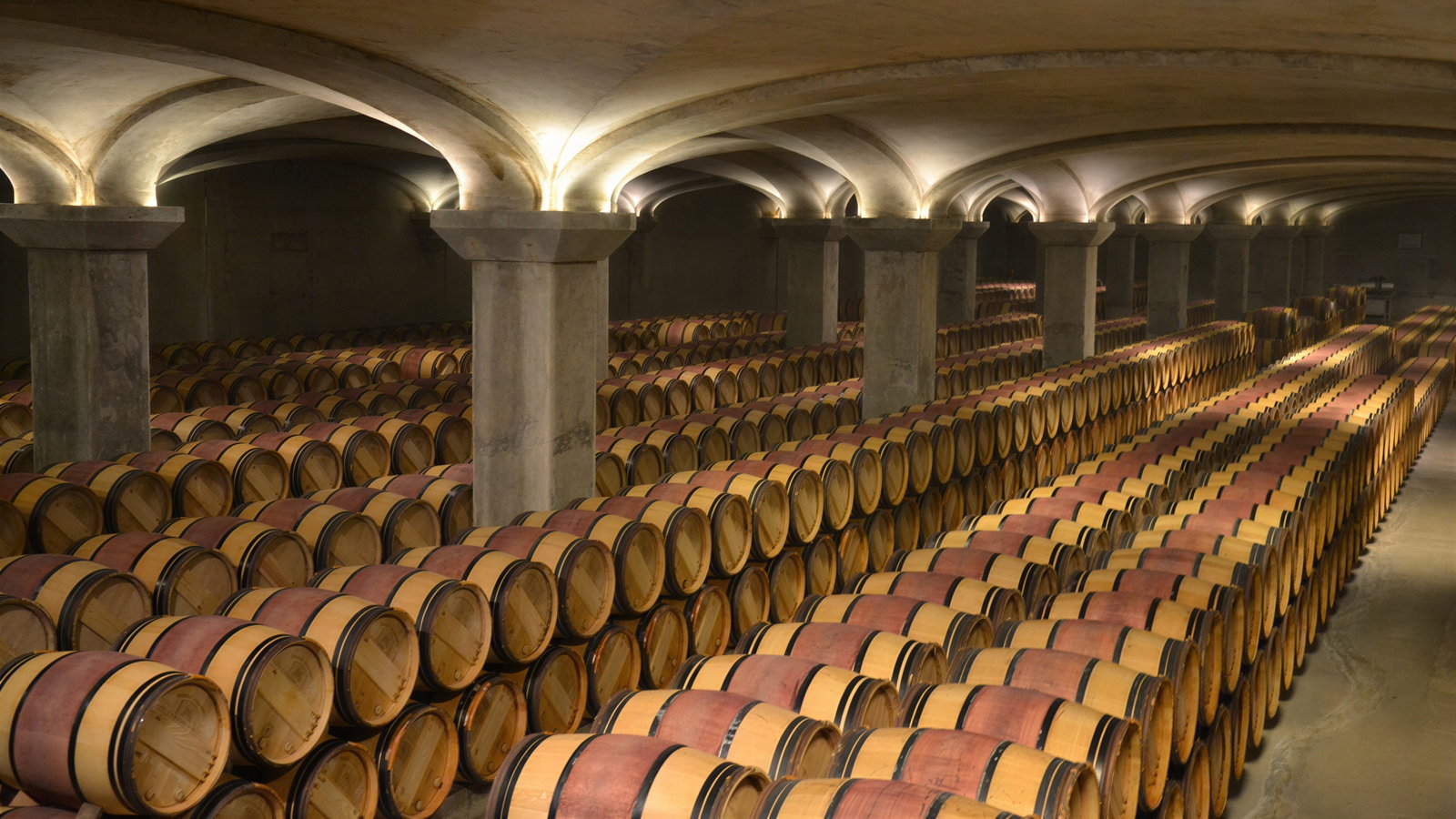 Rows of wine barrels in a dimly lit cellar with arched ceilings and stone pillars, creating a serene and historic atmosphere.