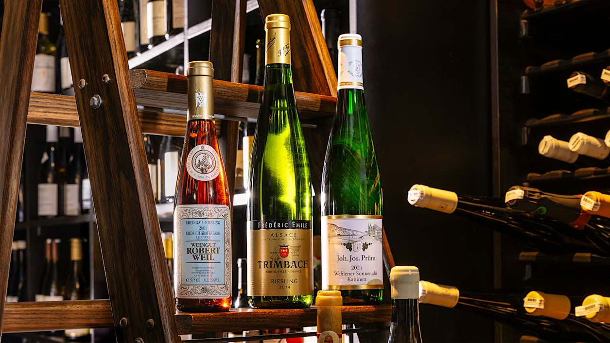 Three wine bottles sit on a wooden rack in a dimly lit wine cellar. Labels show different brands. Background has more wine bottles.