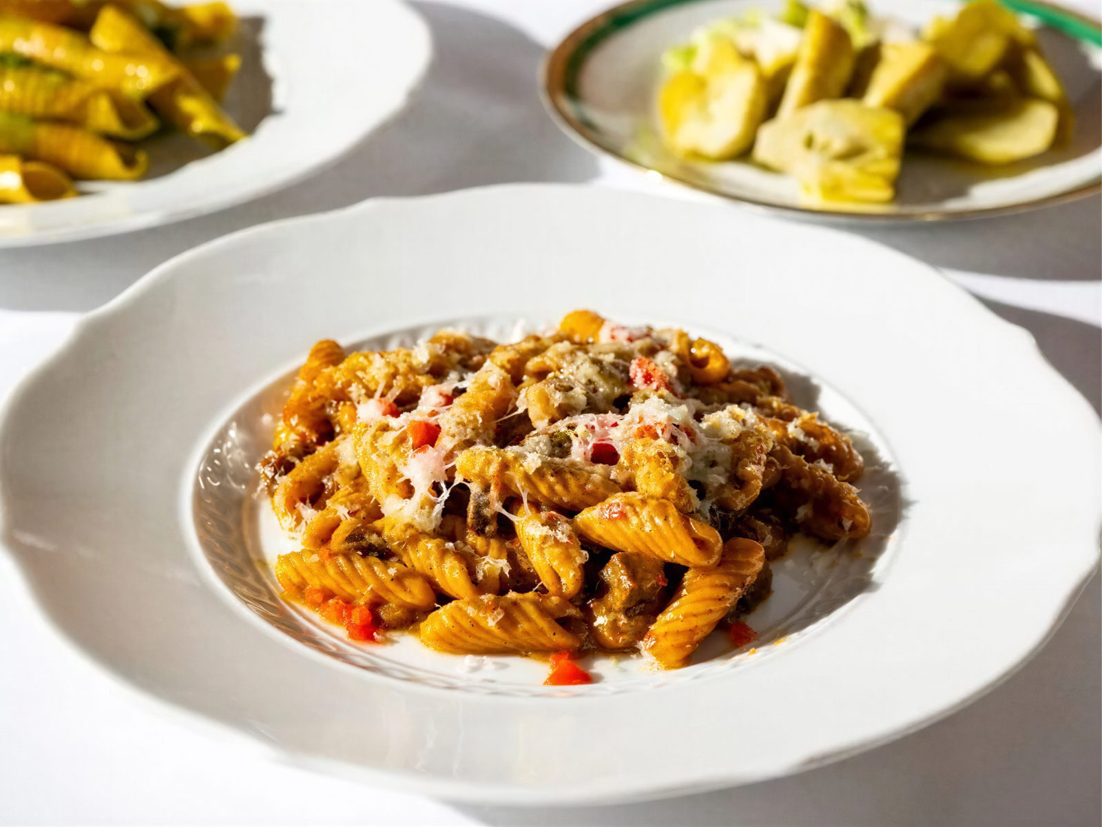 Pasta in a rich sauce topped with cheese on a white plate. Two more dishes with vegetables are in the background on a white tablecloth.
