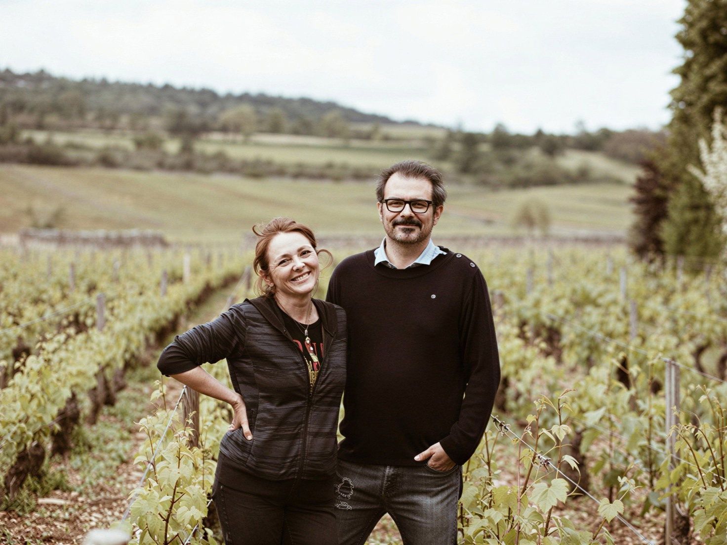 A smiling woman Véronique Bonin and her husband Nicolas Bernard stand in a vineyard, surrounded by lush green grapevines, with rolling hills in the background on a cloudy day.