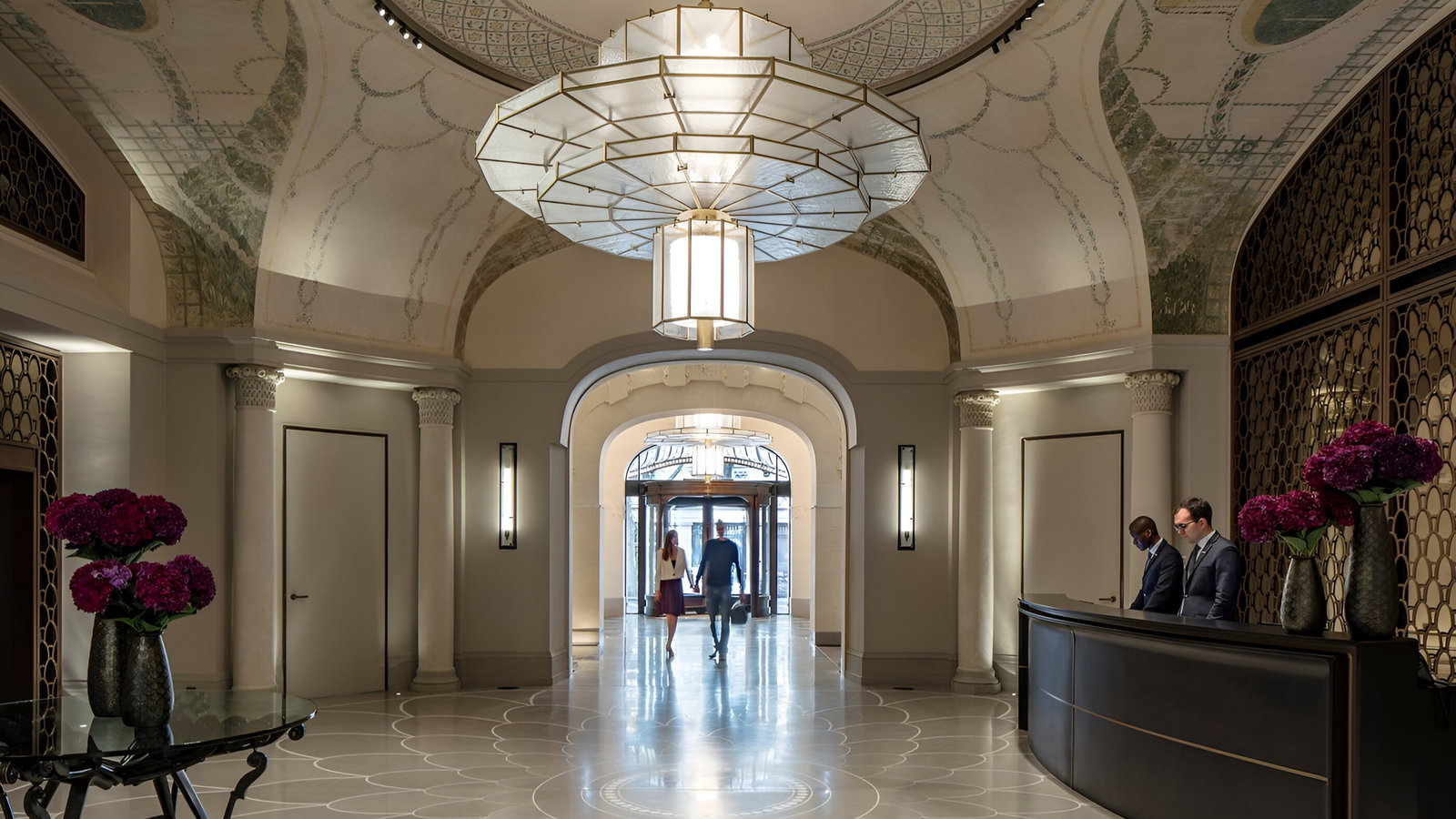 Elegant hotel lobby with ornate ceiling, two staff at reception desk, couple walking toward exit. Purple flowers in vases add color.