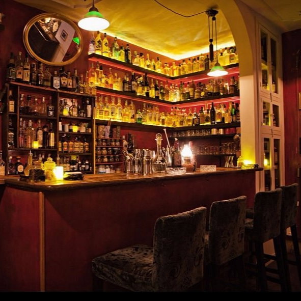 Cozy bar with a warm glow, shelves stocked with bottles, and cushioned stools. Dim lighting and framed photos create a vintage ambiance.