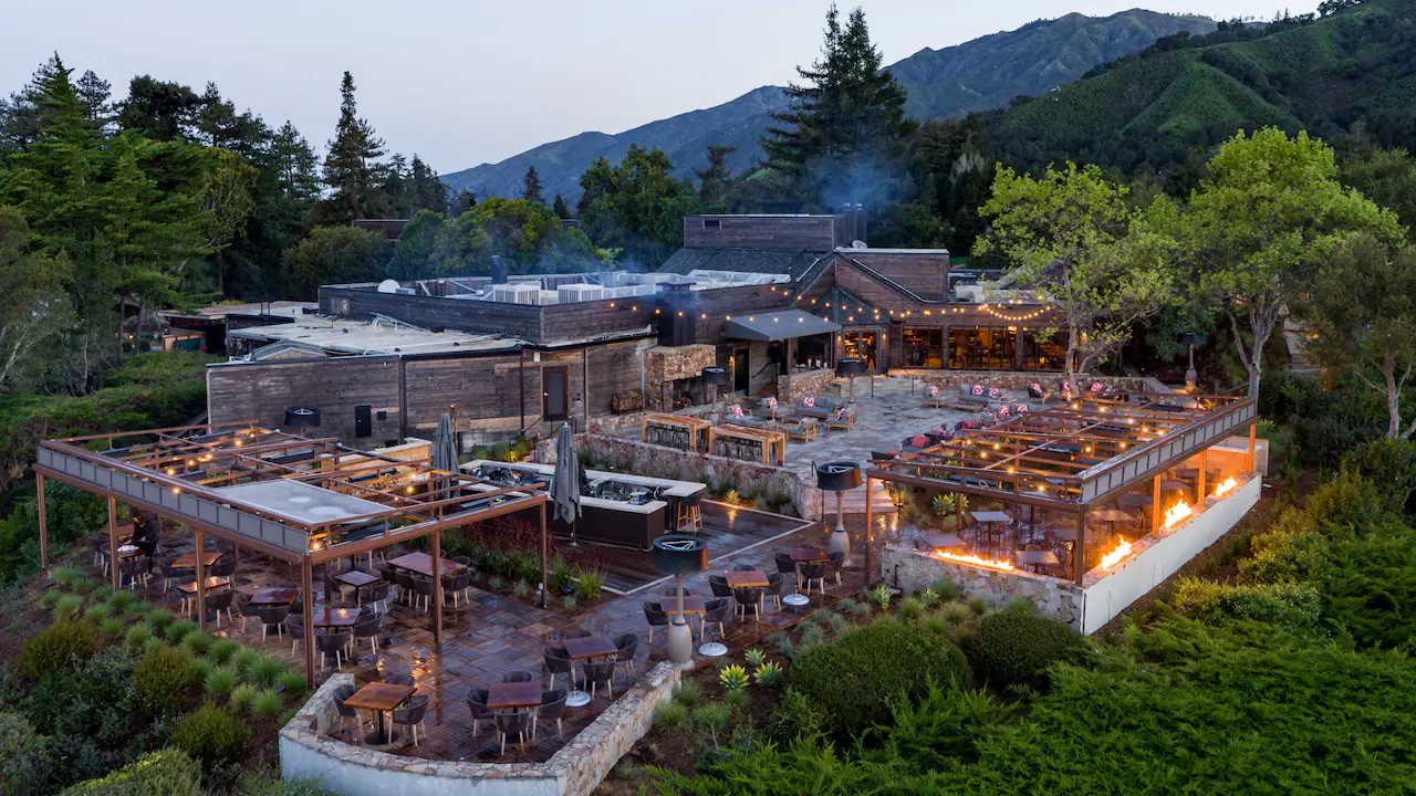Upscale outdoor restaurant with wooden tables, string lights, and fire pits. Surrounded by lush greenery and mountains in the background.