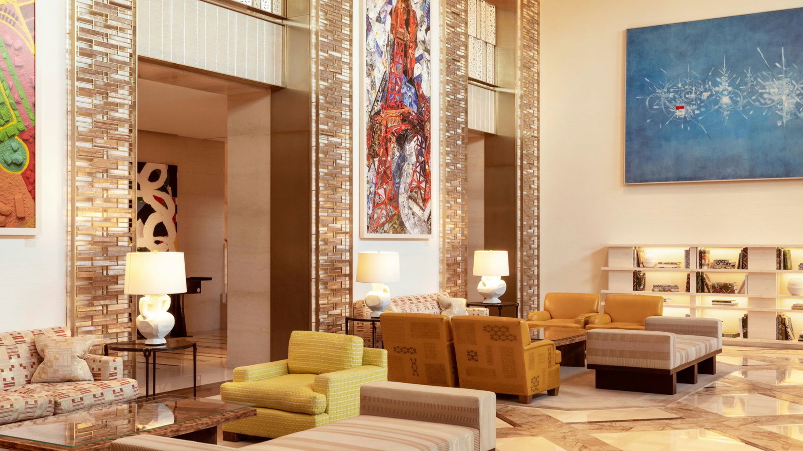 Modern hotel lobby with colorful abstract art, yellow and patterned seating, and warm lighting. Shelves of books add a cozy touch.