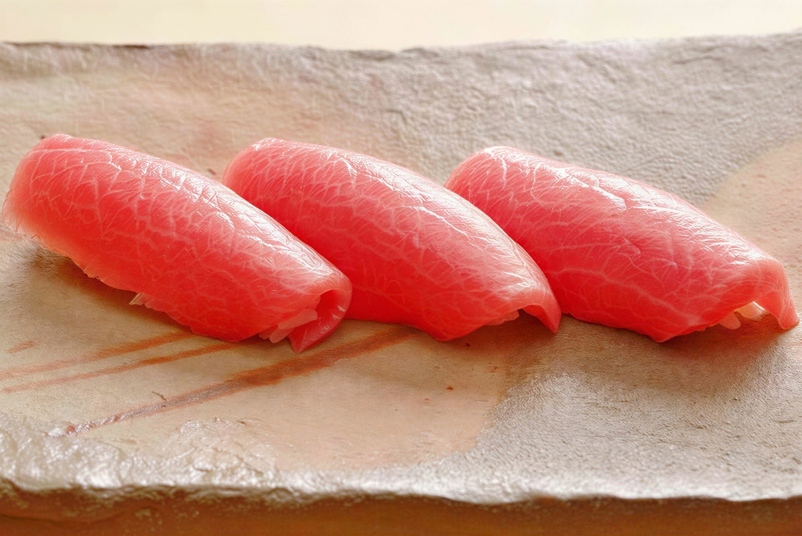 Three pieces of tuna sushi with pink, marbled fish slices on a textured beige plate. The setting conveys a fresh and appetizing mood.