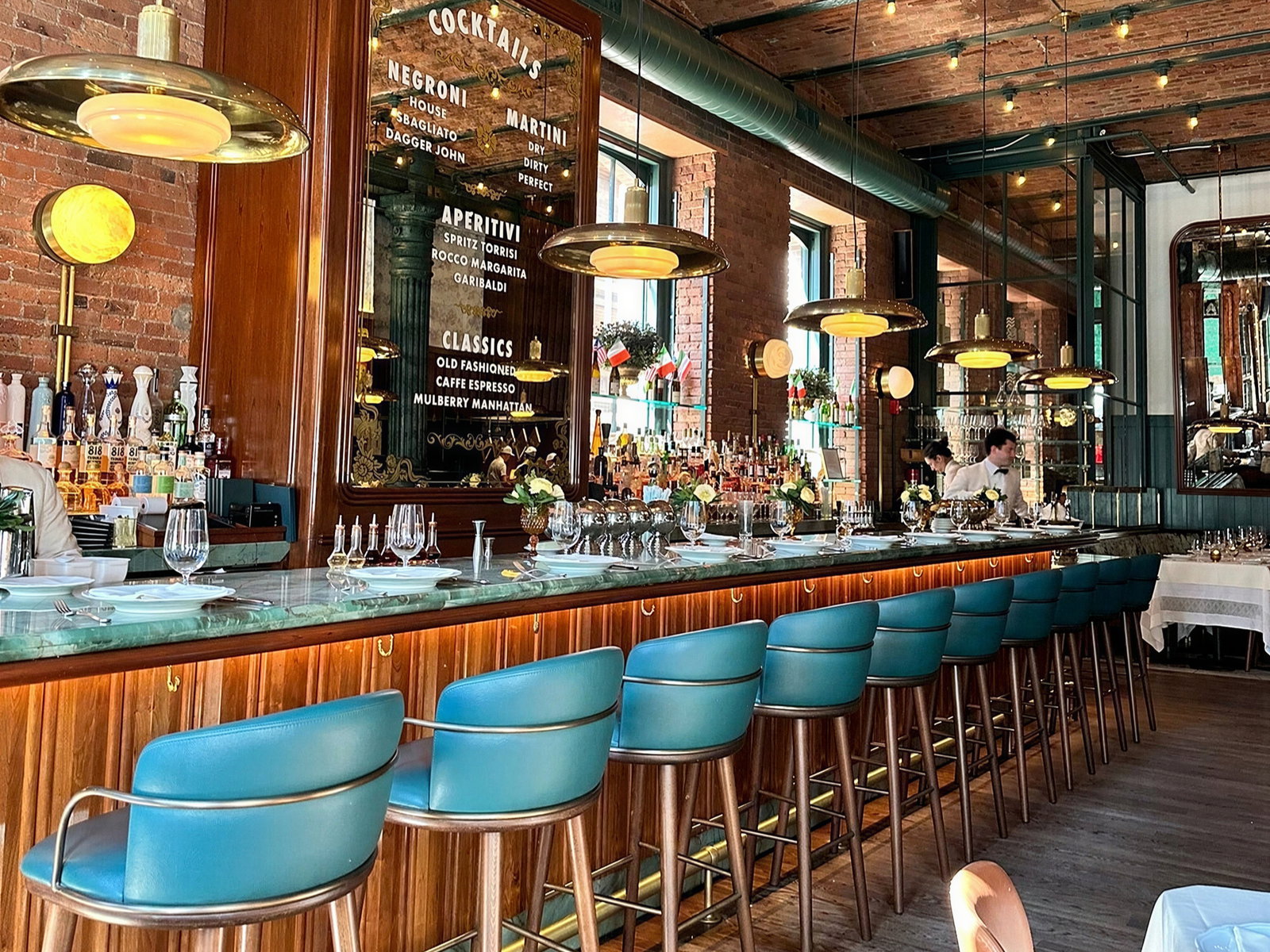 Chic bar with turquoise stools, brick walls, and hanging lights. Bartenders in white shirts. Menu board lists cocktails like Negroni and Martini.
