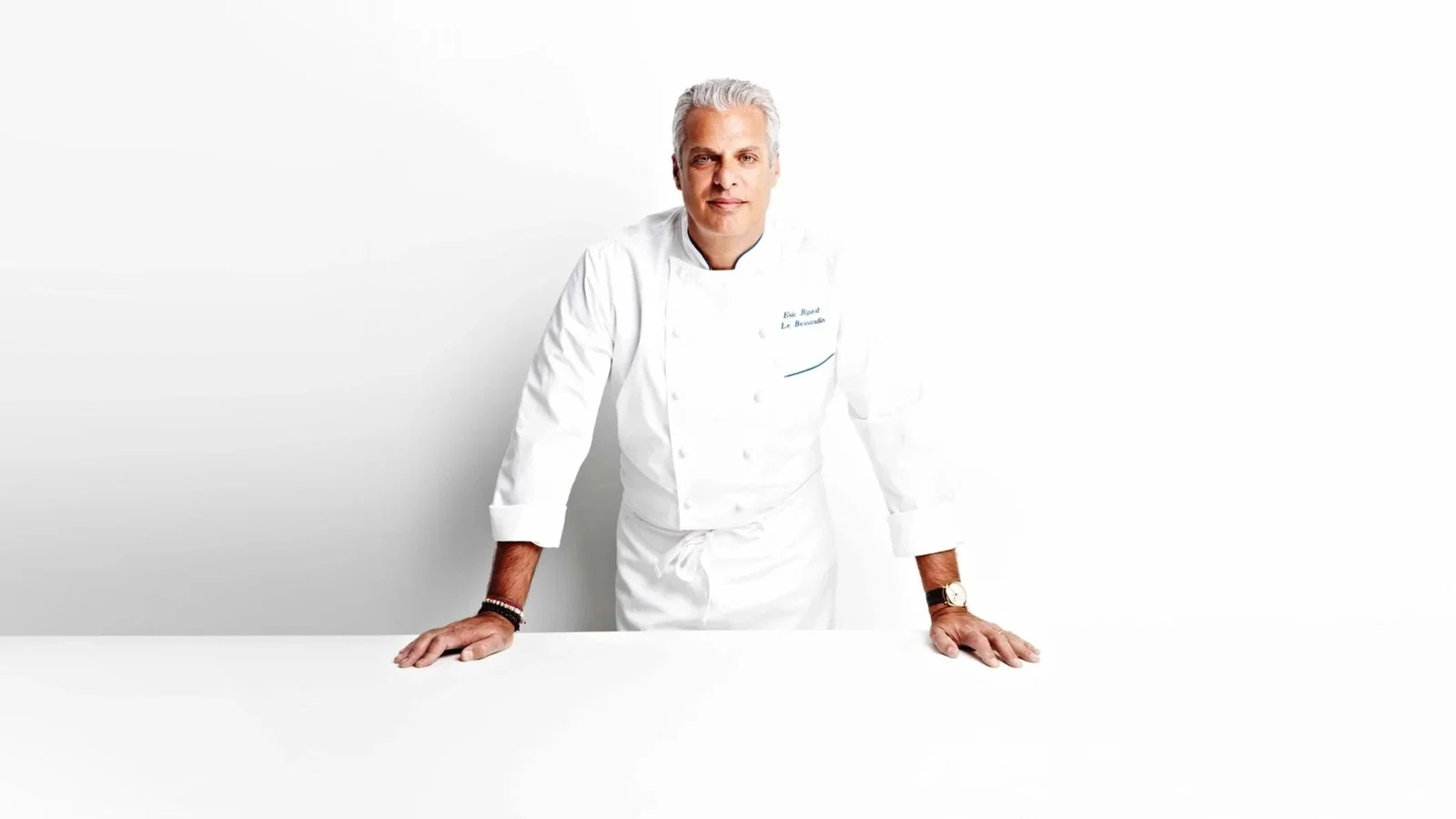Chef in a white uniform leans on a white table against a plain background, exuding confidence. Text on jacket reads: "Eric Ripert, Le Bernardin."