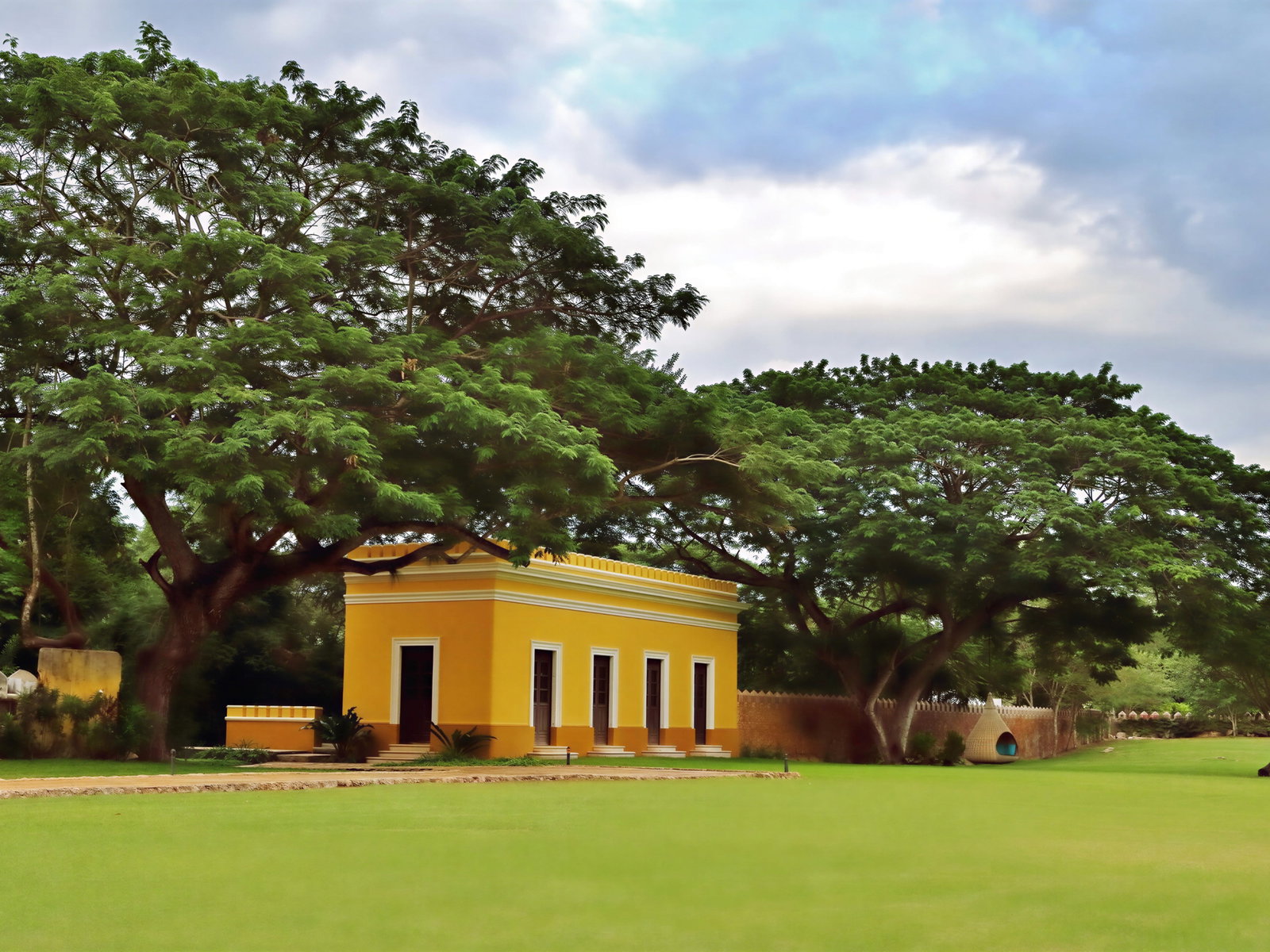 A yellow building with white trim sits among large green trees under a cloudy sky, surrounded by a manicured lawn. The mood is serene.