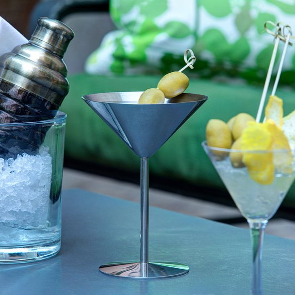 Metal shaker in ice bucket, and two martini glasses with olives and lemon slices on a patio table. Green cushions in the background.