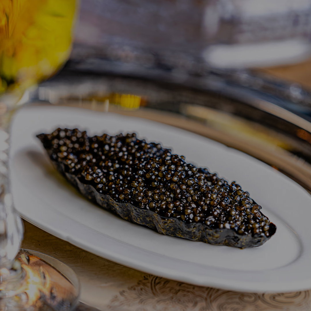 Caviar on a decorative metal tray, served in a ceramic dish. Crystal glass with yellow-green drink nearby adds elegance. Rich, luxurious setting.