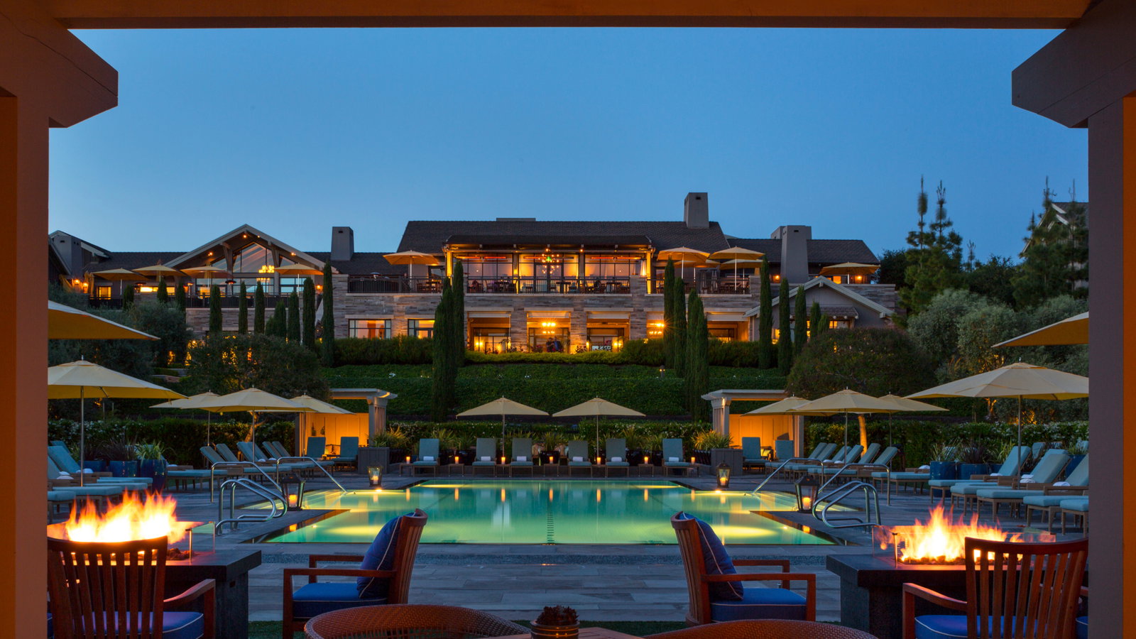 Lit poolside with fire pits and lounge chairs, backed by an illuminated luxury hotel at dusk, creating a serene and inviting ambiance.