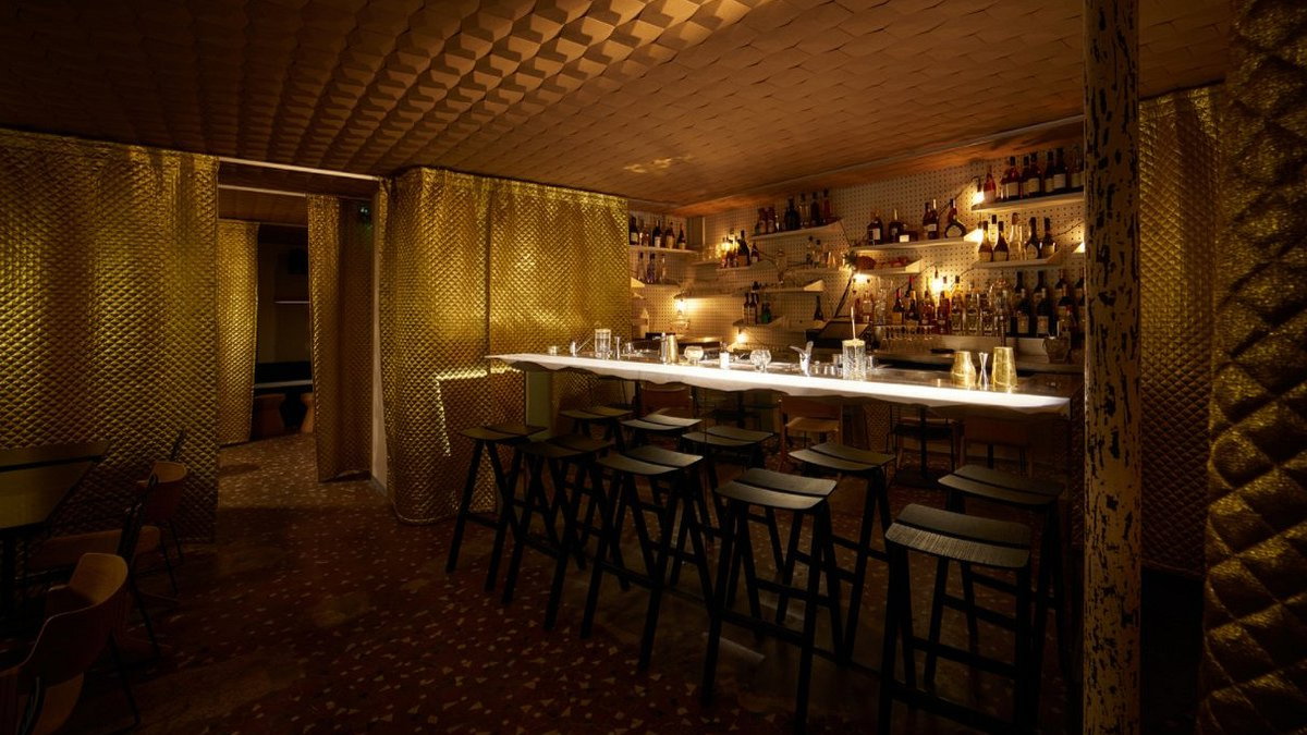 Dimly lit bar with black stools, golden textured curtains, and shelves of bottles. Warm, cozy atmosphere with a modern design.
