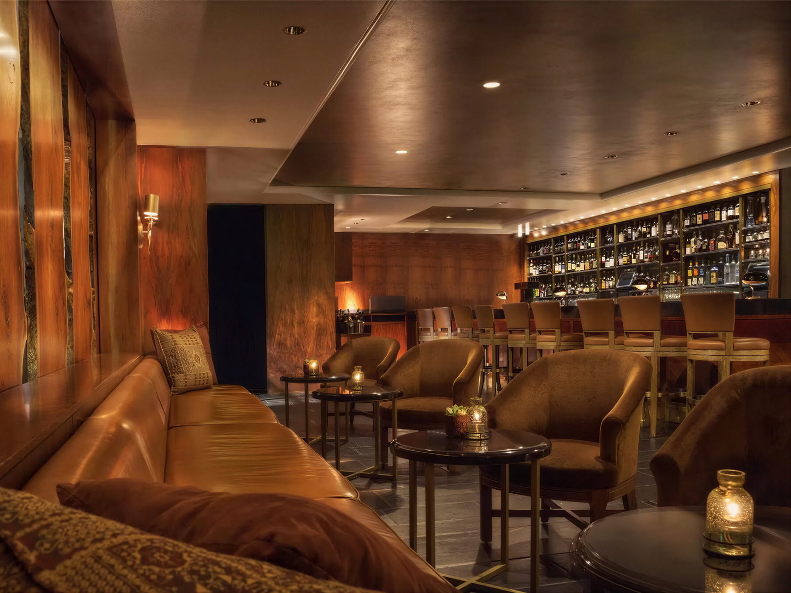 Cozy lounge bar with brown leather sofas and chairs, dim lighting, and a well-stocked bar. Warm wood tones create an inviting atmosphere.