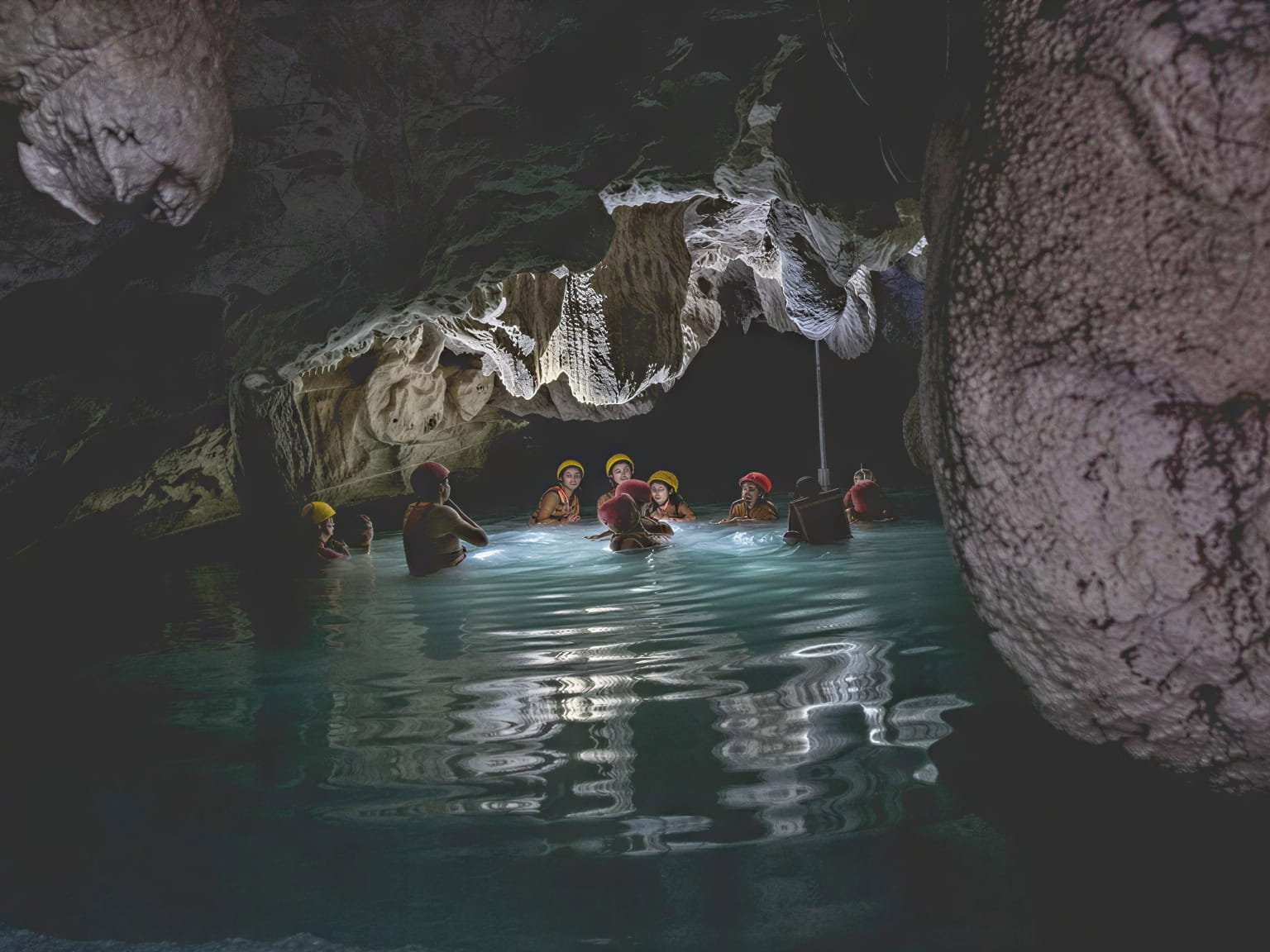 People in helmets swim at Wakax Hacienda in a dimly lit cave with turquoise water, surrounded by stalactites. The atmosphere is adventurous and serene.